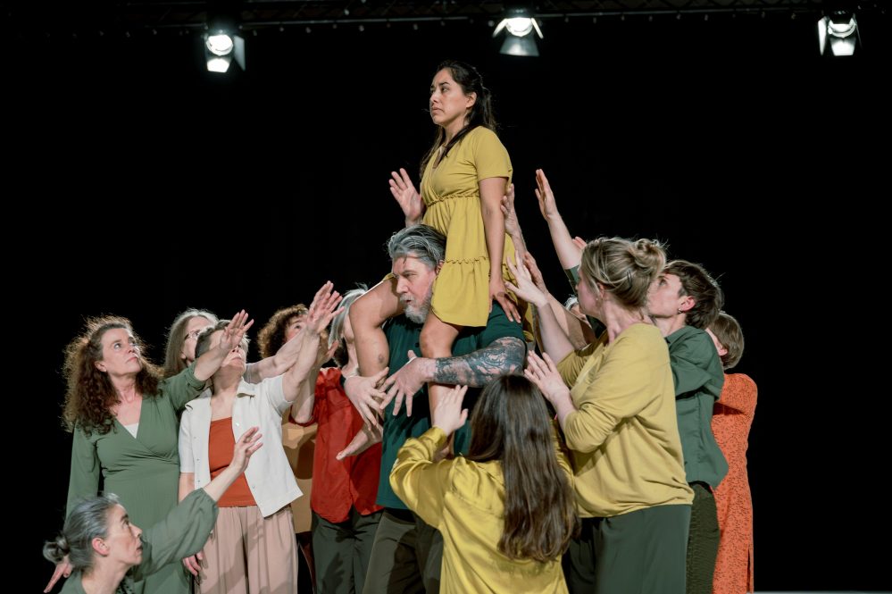 A group of people in various colored outfits lift a woman in a yellow dress overhead. The setting is a stage with black background and overhead lights.