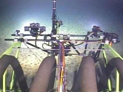 Ein Tiefsee-Bergbau-Roboter sammelt auf dem Meeresboden Manganknollen. Es sind Arme, Kabel und Lampen des Roboters zu sehen, die den Meeresboden voraus beleuchten und durchsuchen.