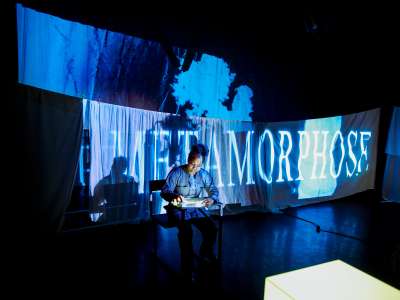 Édith Voges Nana Tchuinang (eine schwarze Frau mit hochgesteckten Haaren in einem grauen Overall) sitzt in einem Bühnenraum vor Licht-Projektionen. EIne der Projektionen schreibt groß "Metamorphose" auf die weißen Tücher, die als Projektionsfläche dienen.