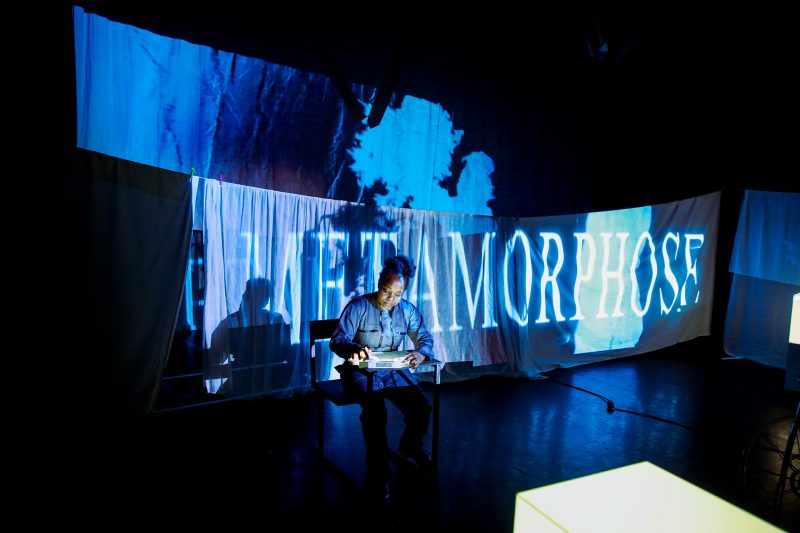 Édith Voges Nana Tchuinang (eine schwarze Frau mit hochgesteckten Haaren in einem grauen Overall) sitzt in einem Bühnenraum vor Licht-Projektionen. EIne der Projektionen schreibt groß "Metamorphose" auf die weißen Tücher, die als Projektionsfläche dienen.