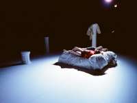 Auf einer dunklen Bühne steht ineinem hellen Lichtkegel ein Bett, in dem zwei Personen liegen. Im Hintergrund stehen mehrere Säulen unterschiedlicher Größe als Kulisse.