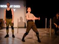 Two people in military attire on stage with a sign saying "Nobody died today"