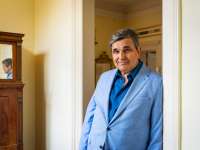 Ein Mann mit dunklen, grau-melierten Haaren, einem hellblauen Jacket und einemblauen Hemd darunter, steht seitlich entspannt angelehnt in einem Türrahmen und blickt freunlich in die Kamera