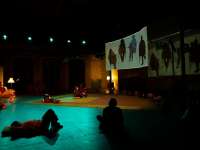 EInige Personen sitzen und liegen in einem Bühnenraum. Sie haben Kopfhörer auf den Ohren und schauen alle in Richtung einer Projektion auf der rechten Seite. Dort sind die Silhouetten von vier Personen zu sehen, die auf Stühlen sitzen. Der Bühnenraum ist mit einem Teppich, ein paar Kissen und eineigen WOhnzimmerlampen ausgestattet, die gemütlicheres Licht ausstrahlen.