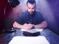 David Kebekus (Mann mit Vollbart und mit langen Haaren, die zum Dutt gebunden sind) sitzt mit einem Stift in der Hand an einem Tisch über einigen Blättern Papier. Neben ihm steht eine Schreibtischlampe. Hinter ihm sind ist Nebel zu sehen. Ein großer Schweiwerfer wirft von hinter David Kebekus Licht auf die Szenerie.