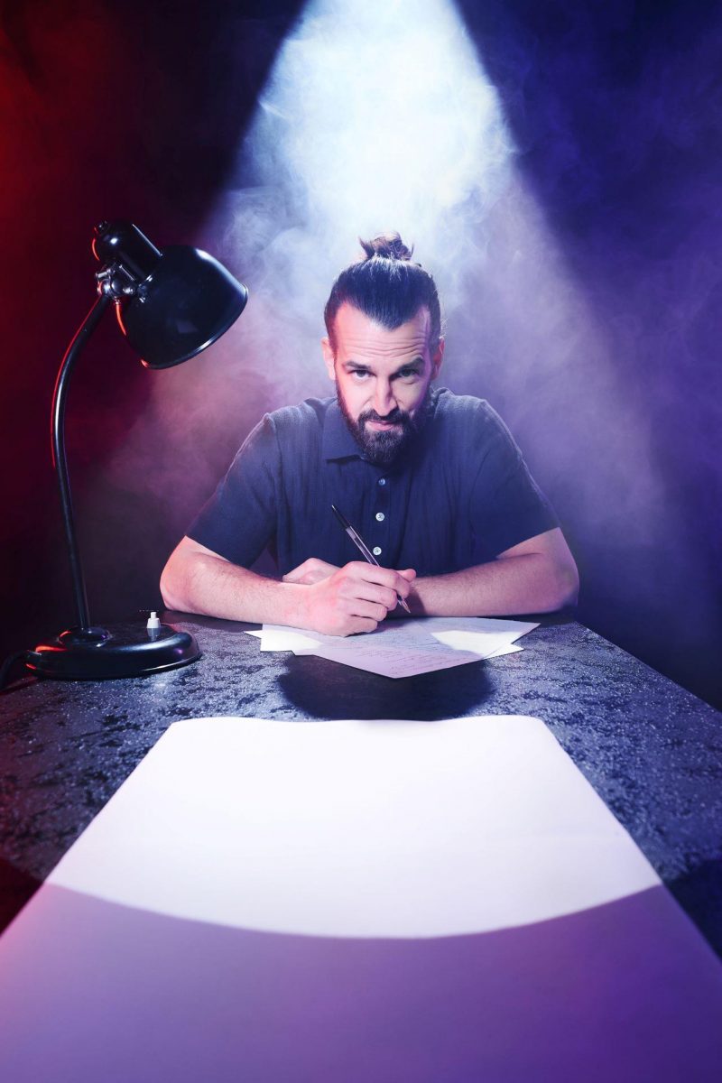 David Kebekus (Mann mit Vollbart und mit langen Haaren, die zum Dutt gebunden sind) sitzt mit einem Stift in der Hand an einem Tisch über einigen Blättern Papier. Neben ihm steht eine Schreibtischlampe. Hinter ihm sind ist Nebel zu sehen. Ein großer Schweiwerfer wirft von hinter David Kebekus Licht auf die Szenerie.