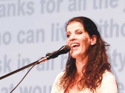 Eine Frau mit braunen, gelockten Haaren, die ihr bis über die Schultern reichen, gekleidet in ein weißes Oberteil, steht an vor einem Mikrofon und singt.