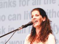 Eine Frau mit braunen, gelockten Haaren, die ihr bis über die Schultern reichen, gekleidet in ein weißes Oberteil, steht an vor einem Mikrofon und singt.