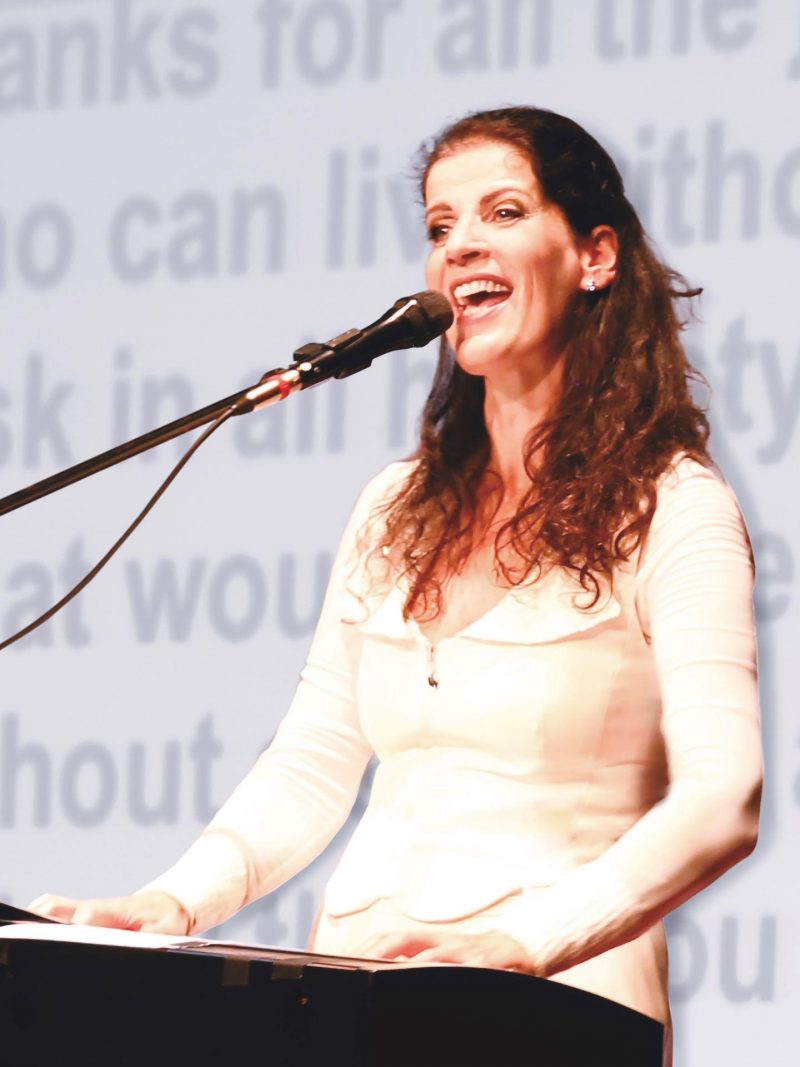 Eine Frau mit braunen, gelockten Haaren, die ihr bis über die Schultern reichen, gekleidet in ein weißes Oberteil, steht an vor einem Mikrofon und singt.