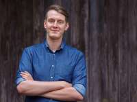 Jonas Greiner (ein Mann mit dunkelblonden Haaren im Kurzharschnitt, blauen Augen und glatt rasiertem Gesicht) steht im blauen Jeanshemd mit verschränkten Armen vor eine Wand aus dunklem Holz und blickt freundlich in die Kamera.