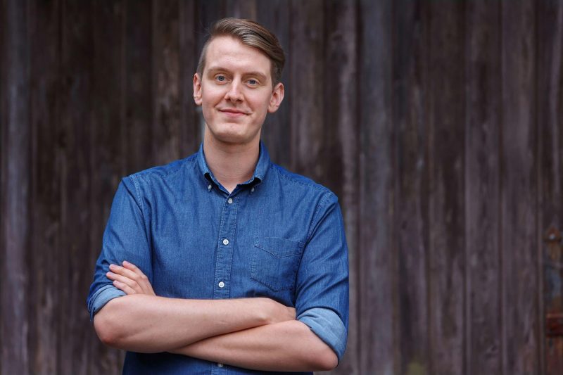 Jonas Greiner (ein Mann mit dunkelblonden Haaren im Kurzharschnitt, blauen Augen und glatt rasiertem Gesicht) steht im blauen Jeanshemd mit verschränkten Armen vor eine Wand aus dunklem Holz und blickt freundlich in die Kamera.