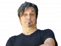 Muhsin Omurca hat dunkle, leicht gräuliche Haare und einen Kinnbart. Er trägt ein schwarzes T-Shirt und zwinkert leicht von oben herab in die Kamera.