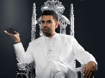 Tan Caglar (ein Mann Mitte 40, mit dunklen Haaren, die im Kurzhaarschnitt mit Gel gestyled sind, einem gepflegten Dreitagebart) sitzt in einem weißen Anzug auf einem silbernen Thron vor einem dunklen Hintergrund. Auf seiner rechten Hand präsentiert er einen kleinen Rollstuhl. Mit strengem Blick schaut Tan Caglar in die Kamera.