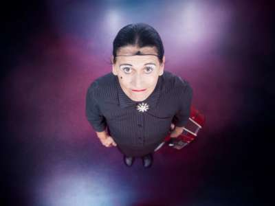 Carlema De Feo ist von oben fotografiert zu sehen. Sie hebt den Kopf (mit Haarnetz) und blickt nach oben in die Kamera. Der Kopf ist durch die Perspektive nah an der Kamera und größer als der Körper. Ihr Akkordeon steht neben ihr auf dem Boden.