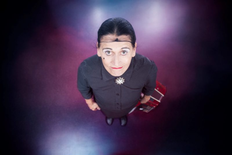 Carlema De Feo ist von oben fotografiert zu sehen. Sie hebt den Kopf (mit Haarnetz) und blickt nach oben in die Kamera. Der Kopf ist durch die Perspektive nah an der Kamera und größer als der Körper. Ihr Akkordeon steht neben ihr auf dem Boden.