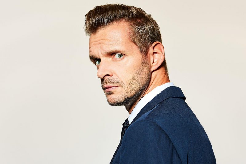 Florian Schroeder schaut über seine Schulter in die Kamera. Er trägt einen blauen Anzug und darunter ein weißes Hemd. Er ist weiß, hat blaue Augen, blondbraune Haare und einen 3-Tage-Bart.