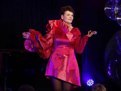 Gayle Tufts steht im roten Kleid, mit Mikrofon in der Hand vor dunklem Hintergrund auf einer Bühne. Disco-Kugeln schmücken den Hintergrund.