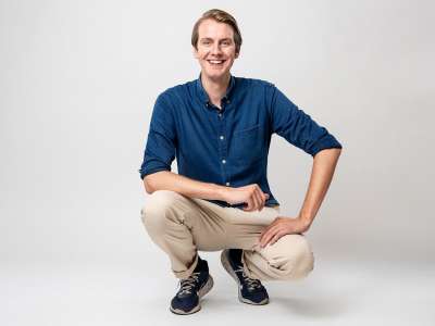 Jonas Greiner (ein Mann in blauem Jeanshemd und beiger Hose und blauen Turnschuhen mit dunkelblonden Haaren, die locker zur Seite gekämmt sind) hockt vor einem weißem Hintergrund und schaut strahlend lächelnd in die Kamera.
