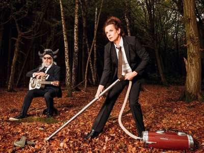 Nessi Tausenschön (eine Frau mit braunen Haaren in einem losen Dutt, die einen schwarzen Anzug, ein weißes Hemd und eine Kravatte trägt) steht in einem Wald und staubsaugt mit einem Retro-Staubsauger den Waldboden voller Laub. Ein Mann mit langen grauen Haaren, grauem Bart, SOnnenbrille und einer Mütz mit Plüsch-Kuhhörnern sitzt mit einer Gitarre schräg hinter ihr im Wald auf einem Hocker.