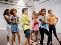 Eine Gruppe von Kindern und Jugendlichen steht in einem hellen Raum mit weißen Wänden. Alle machen eine coole Pose mit einer Hand am Kinn.