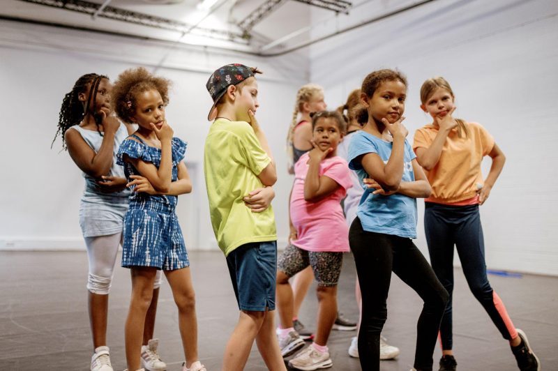 Eine Gruppe von Kindern und Jugendlichen steht in einem hellen Raum mit weißen Wänden. Alle machen eine coole Pose mit einer Hand am Kinn.