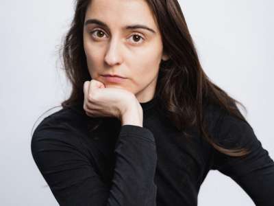 Anna Heithausen (eine junge Frau mit braunen Haaren und braunen Augen, trägt einen schwarzen Pullover) schaut in die Kamera. Dabei stützt sie den Kopf auf einem Arm ab.