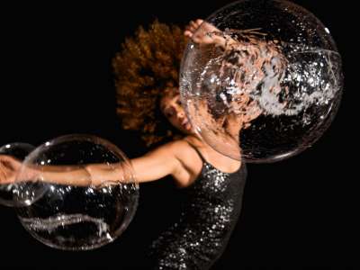 Ein*e Schwarze Performer*in in dynamischer Pose mit wehenden Locken, einem seitlich ausgestreckten Arm, dem zweiten Arm halb über dem Kopf erhoben. und glitzerndem Klei ist vor dunklem Hitergrund verschwommen zu sehen. durchsichtige, aber strukturierte Kugeln, wie Seifenblasen scheinen an den Armen zu hängen und den Blick auf die Person zu verzerren.