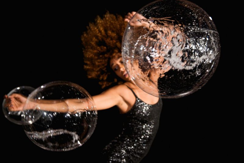 Ein*e Schwarze Performer*in in dynamischer Pose mit wehenden Locken, einem seitlich ausgestreckten Arm, dem zweiten Arm halb über dem Kopf erhoben. und glitzerndem Klei ist vor dunklem Hitergrund verschwommen zu sehen. durchsichtige, aber strukturierte Kugeln, wie Seifenblasen scheinen an den Armen zu hängen und den Blick auf die Person zu verzerren.