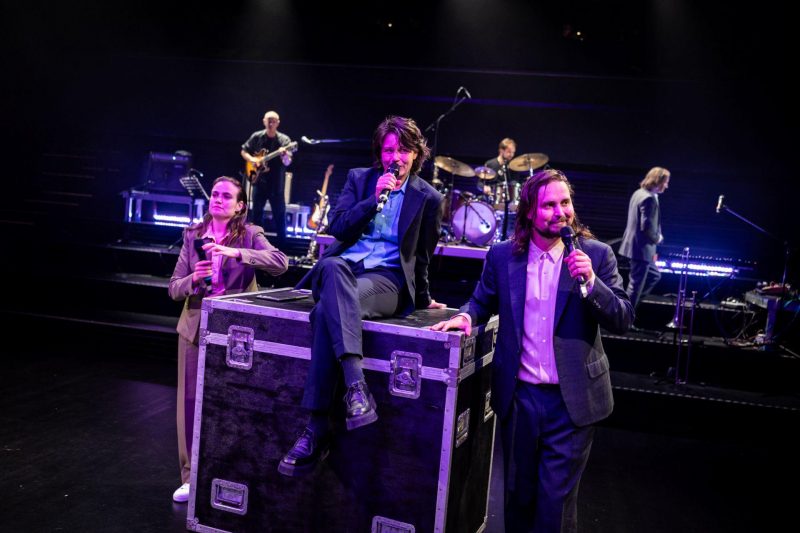 Drei Personen in Anzügen und Hemden sind im Bildvordergrund. Zwei stehen rechts und links eines großen Flightcase (Koffers), die dritte Person sitzt mit überschlagenen Beinen darauf.Alle haben Mikrofone in der Hand. Im Hindergrund ist eine Band zu sehen.