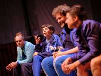 Vier Schauspieler*innen sitzen nebeneinander auf einer Bühne, alle tragen blaue Kleidung, eine Person spricht gestikulierend