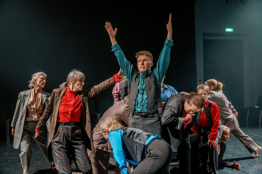 Gruppe von Menschen in unterschiedlicher Kleidung in einer dynamischen Tanzpose auf einer Bühne mit dunklem Hintergrund