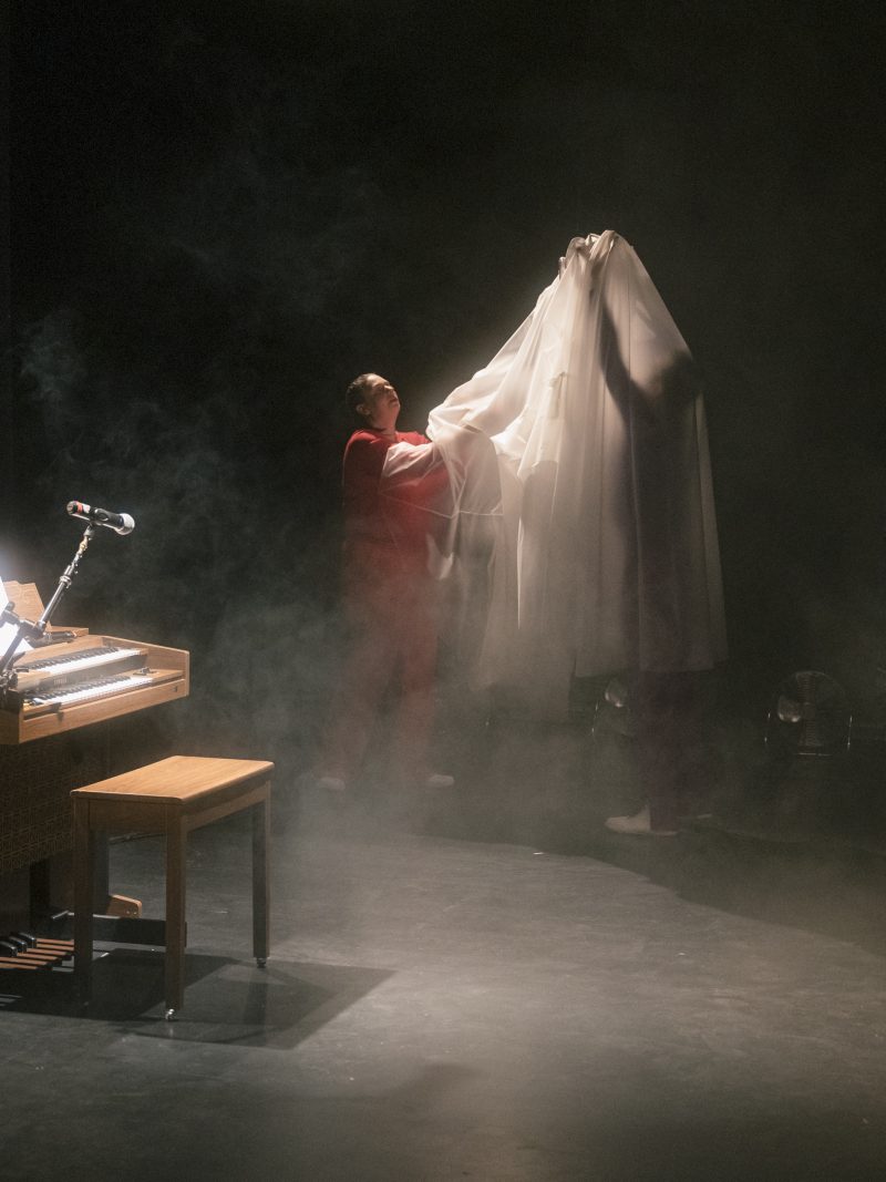 Person hebt ein großes, transparentes Tuch auf einer Bühne mit Nebel neben einem Klavier mit Mikrofon