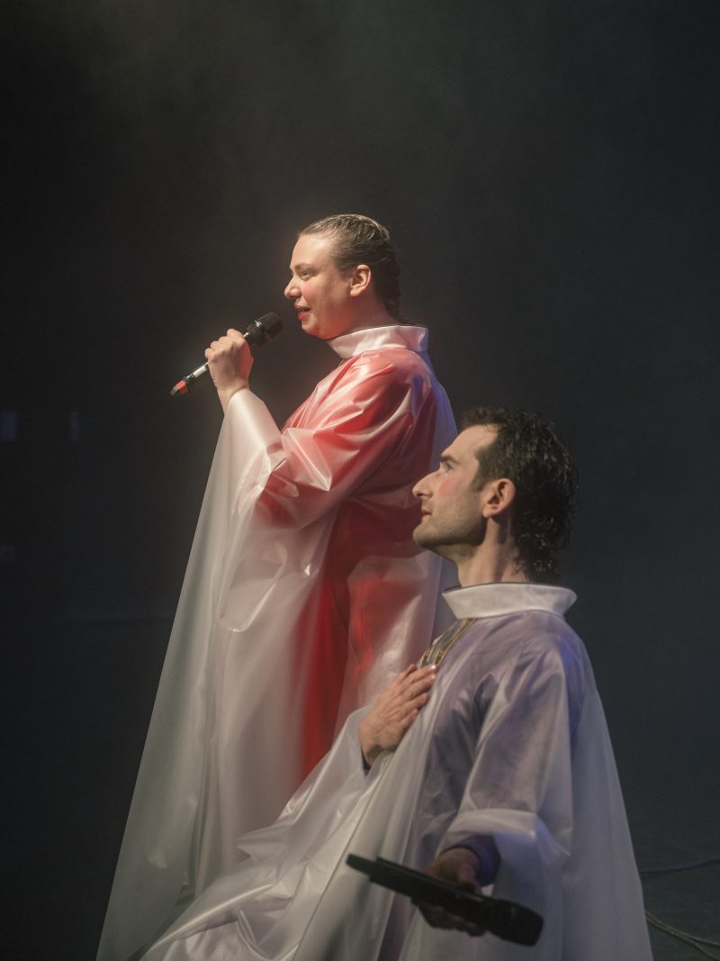 Zwei Personen in durchsichtigen Regenponchos auf einer Bühne, eine steht mit Mikrofon, die andere sitzt mit Mikrofon in der Hand