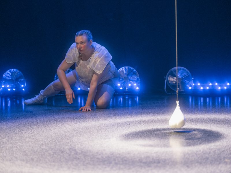 Person in futuristischer Kleidung kniet auf einem sandbedeckten Boden vor einem Loch, aus dem ein an einem Seil hängendes Objekt herabfällt