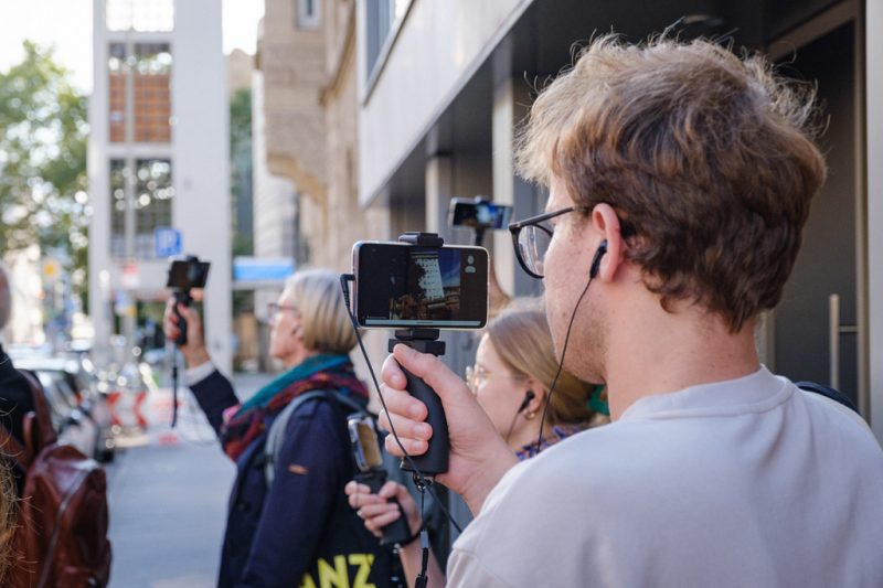 Es sind mehrere Personen zu sehen. Sie stehen in einer Straße neben einem Gebäude und halten Smartphones in der Hand auf denen eine andere Stadt zu sehen ist. Die Personen haben auch Kabelkopfhörer in den Ohren, deren Stecker in das Smartphone eingesteckt sind.