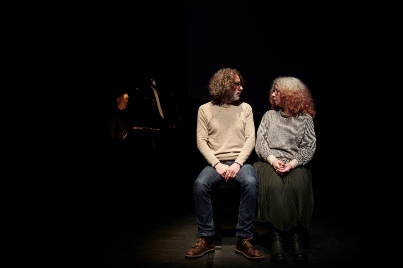 Zwei Personen sitzen in einem dunklen Raum eng nebeneinander. Sie werden mit einem Spotlight beleuchtet. Ihre Köpfe sind einander zugewandt und sie schauen sich gegenseitig an.