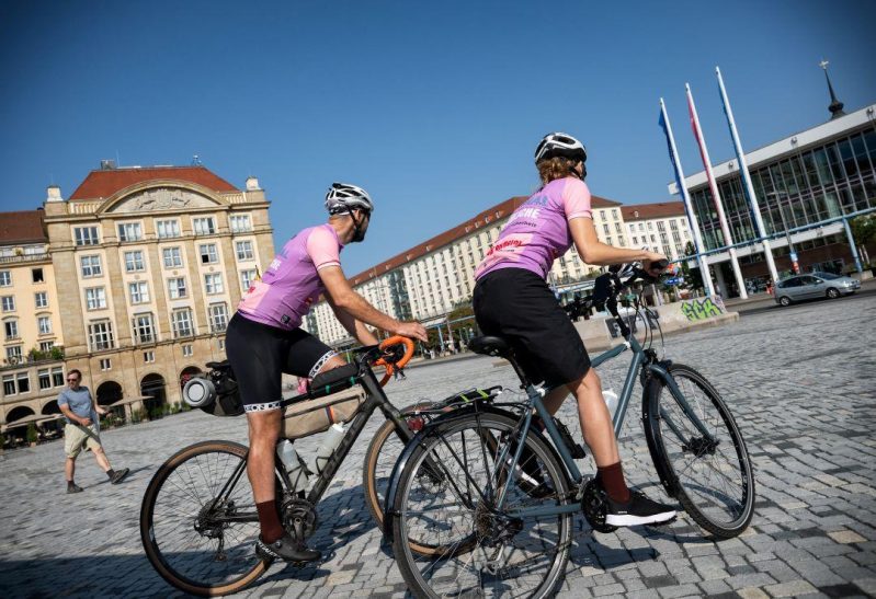 Zwei Menschen in rosfarbenen Fahrradtrikots und mit Fahrradhelmen auf fahren bei gutem Wetter über einen großen gepflasterten Platz.