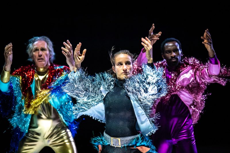 Drei Menschen in bunten Kostümen stehen auf der Bühne und heben ihre Hände.