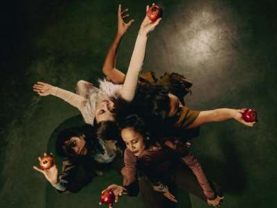 Vier weiblich gelesene Performer*innen sind aus der Vogelperspektive zu sehen. Sie hocken Rücken an Rücken. Alle von ihnen halten einen roten Apfel im ausgestreckten Arm - den Blick jeweils fest auf ihren Apfel gerichtet.