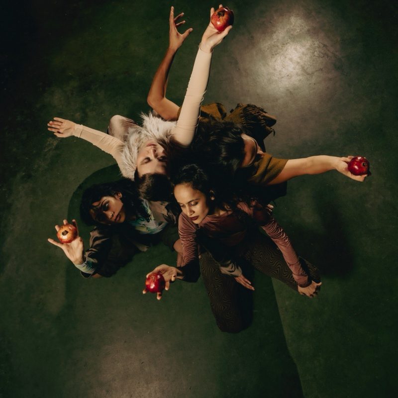 Vier weiblich gelesene Performer*innen sind aus der Vogelperspektive zu sehen. Sie hocken Rücken an Rücken. Alle von ihnen halten einen roten Apfel im ausgestreckten Arm - den Blick jeweils fest auf ihren Apfel gerichtet.