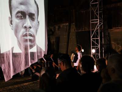 Eine zeitgenössische Live-Performance mit einem Musiker, der auf der Bühne neben einer großen hängenden Projektionsfläche, auf der ein schwarz-weißes Porträtgesicht zu sehen ist, Akkordeon spielt; das Publikum sitzt in einem schwach beleuchteten Industriegebäude.