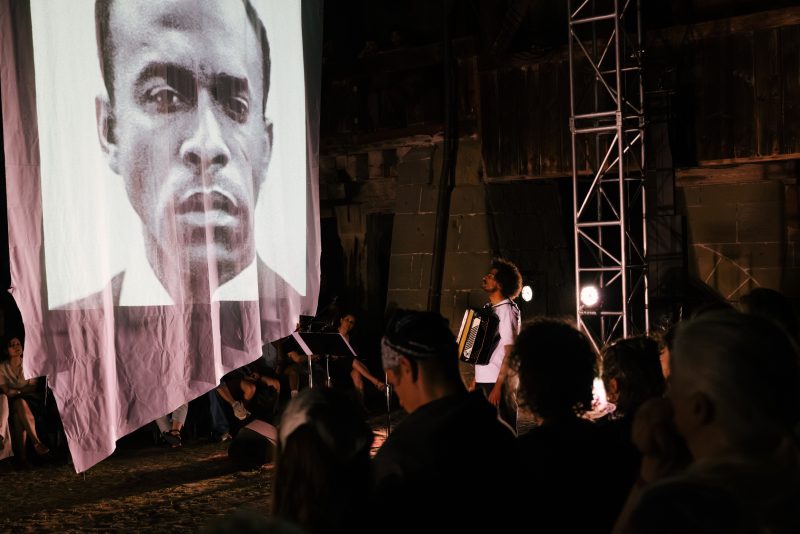 Eine zeitgenössische Live-Performance mit einem Musiker, der auf der Bühne neben einer großen hängenden Projektionsfläche, auf der ein schwarz-weißes Porträtgesicht zu sehen ist, Akkordeon spielt; das Publikum sitzt in einem schwach beleuchteten Industriegebäude.