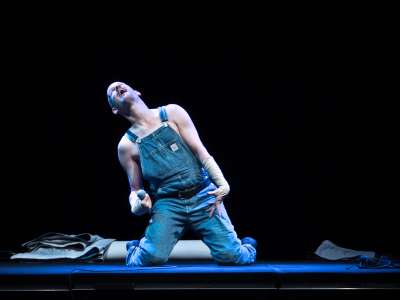 Eine Person mit Glatze und Latzhose kniet auf einer Bühne mit zurückgeworfenem Kopf und ausgebreiteten Armen.