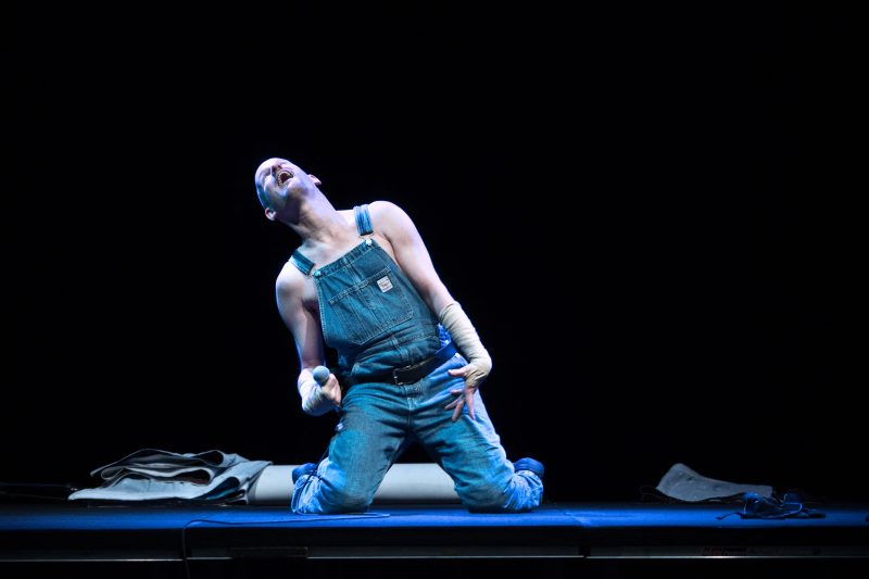 Eine Person mit Glatze und Latzhose kniet auf einer Bühne mit zurückgeworfenem Kopf und ausgebreiteten Armen.