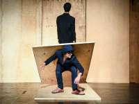 Ein Mann in dunkler Kleidung hockt mittig auf einer Bühne. Auf seinen Schultern liegt eine Holzplatte, die nach hinten zum Boden hin abfällt. Dahinter steht ein weiterer Mann, auch in dunkler Kleidung, mit dem Rücken zu uns vor einer Wand aus ähnlichen Holzplatten, wie die die auf den Schultern des hockenden Mannes aufliegt.