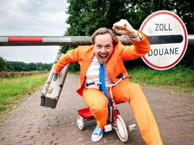 Patrick Nederkoorn (weißer Mann mit blonden Haaren und Drei-Tage-Bart) sitzt in in knallig-orangenem Anzug mit weißem Hemd, blauer Krawatte und einem alten Koffer in der Hand auf einem kleinen Dreirad und scheint gerade unter einer Schranke mit Zoll-Schild druchgefahren zu sein. Sein Gesicht ist freudestrahlend und seine Gesten überschwänglich. Der Hintergrund des Bildes sind grüne Laubbäume und die kleine gepflasterte Straße zu sehen auf der Nederkoorn gekommen zu sein scheint.