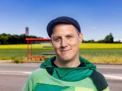 Sebastian 23 (Mann mit grünem Hoodie und schwarzer Schiebermütze) vor einer Landstraße mit roter Bushaltestelle und Feldern