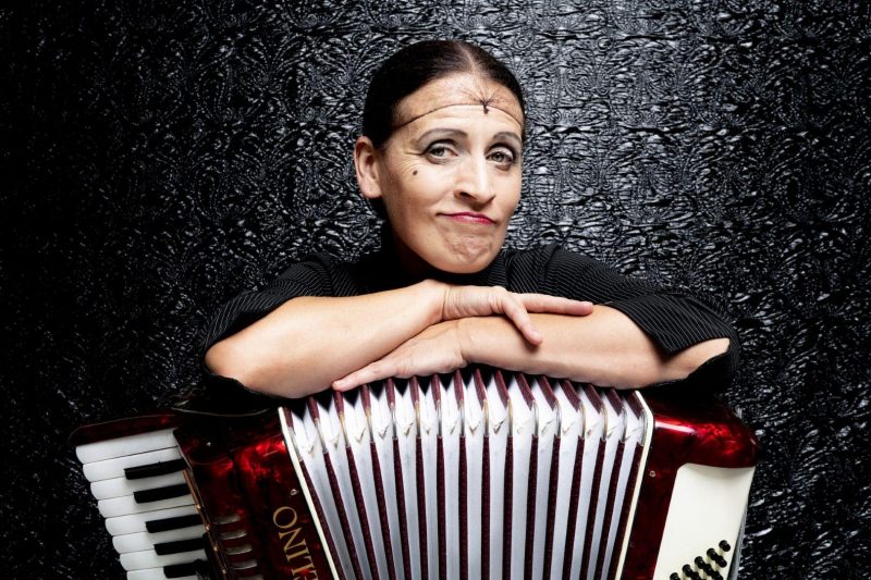 Carmela de Feo (Frau mit dunklen Haaren unter einem Haarnetz, rot geschminkten Lippen und smokey Eyes) lehnt mit verschränkten Armen und einem verschmitzt-herausfordernden Blick auf ihrem roten Akkordeon. Hinter Ihr eine schwarze Wand mit einem im Licht glänzenden Muster.